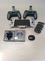 Full view of two black joystick switch controllers, transparent joystick phone controller, Monster N-Lite 203 Bluetooth 5.3 HiFi earbuds with box, and three sets of generic wireless earbuds with charging cases on tabletop.