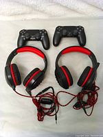 Two black PS4 controllers placed side by side above two pairs of black and red wired headphones with cables coiled on a white cloth background.