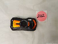 Front view of RICOH WG-70 digital camera showing orange and black rugged body with lens and LED ring light.