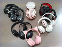 Eight pairs of wireless over-ear headphones on a table: two black Cowin E7 Pro headphones, one pink cat ear headphones, two white pairs, and three other black pairs. All tested and in great condition.