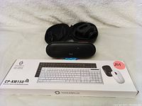 Photo showing Coolerplus CP-KM150 wireless keyboard in original box with Tribit speaker and Soundcore headphones in soft case behind it.