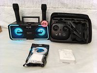 Karaoke machine with blue LED speakers and two microphones next to a black drone with remote control inside a carrying case, alongside a USB C charging cable and a small white box.