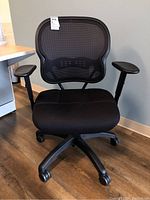 Front view of a black office chair with mesh back, padded seat, and labeling on upper left of the backrest showing item and lot details.