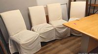 Four muslin slipcovered chairs arranged against wall and table corner on wood floor.