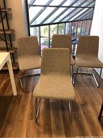 Front angle view of four cantilever chairs with grey fabric seats and backs on chrome metal bases arranged in a room with wood flooring and large window.