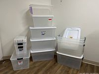 Photo showing 12 stacked plastic storage bins of various sizes, mostly clear with white lids, some with stickers and scratches.