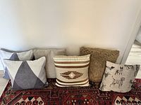 Photo showing six square accent pillows in various neutral shades and patterns arranged on a decorative rug against a white wall.