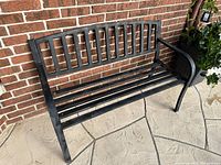 Black metal outdoor bench against a brick wall, showing slatted seat and backrest with armrests on both sides.