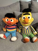 Photo of two large Bert and Ernie plush dolls seated on a couch, showing their full body and detailed features.
