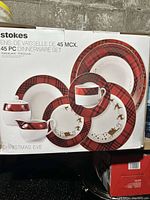 Photo of the box showing Stokes 45 piece Christmas Eve porcelain dinnerware set, featuring red plaid and gold festive details.