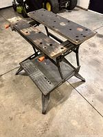 Front angle view of collapsible work table showing metal frame, wooden plank tops with holes and orange adjustment handles.