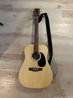 Full frontal image of an acoustic guitar lying on light-coloured wooden floor, showing natural wood finish, black pickguard, fretboard with dot inlays and attached black strap.