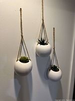 Three white round ceramic hanging plant holders with jute rope hangers on wall, each containing a different artificial succulent plant.