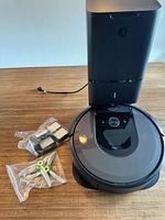 Roomba robot vacuum on wooden surface next to Clean Base station and plastic bags of replacement parts