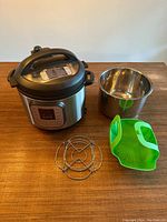 Photo of Instant Pot base unit, stainless steel inner cooking pot, metal steamer rack and green silicone steamer basket on wooden table.