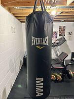 Front view of black Everlast MMA punching bag hanging from ceiling in home gym with some gym equipment in background.