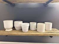 Photo of six white ceramic indoor planters on a wooden shelf against a dark gray wall
