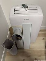 White portable air conditioner unit with Noma branding, remote control on top, plus vent hose parts laid out beside it.