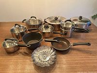 Photo showing a set of nine pots and pans including various sizes of stainless steel pots with lids, two frying pans, and a collapsible steamer basket arranged on a wooden surface.