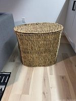 Full view of closed oval rattan laundry basket on light wood floor near sofa and cabinets.