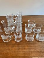 Set of twelve glasses on table. Six mugs with handles and six old-fashioned glasses without handles. Each glassware piece has a frosted sailboat motif etched around.