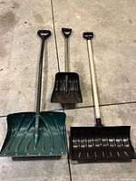 Three plastic snow shovels of varying sizes and colors laid on concrete floor.