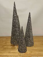 Three cone-shaped silver glitter decorative trees in small, medium, and large sizes shown side by side on a wooden surface against a plain background.