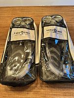 Two black Aqua Lung snorkeling sets in transparent carrying bags, each containing flippers, a mask, and snorkel, placed side by side on a wooden surface.