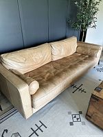 Front-left angle view of tan leather sofa with tufted seat cushion and two cylindrical bolster pillows