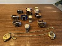 Photo showing all the watches (11 wristwatches) spread on a wooden surface along with 2 gold-tone pocket watch style pieces with chains below them, displaying various strap types and face designs.
