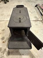 Front view of antique cast iron mini wood stove showing open front door and embossed branding on bottom flap.