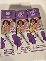 Photo of three packaged M. Rosenfeld Home sink drains showing one gold drain with overflow and two brushed nickel drains without overflow in original purple and white boxes.