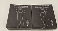 Two unopened boxed water leak detectors with black packaging and sensor alarm graphics, side by side