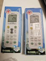 Two K-1028V universal air conditioner remote controls in their original blue and white packaging boxes. LCD display and multiple buttons visible through the package design.