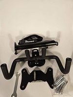 Top-down view of black bike wall mount hanger with foldable hooks, mounting bracket, four metal expansion screws, an M5 hex wrench, a small open-end wrench, and a small carry handle strap labeled 'Deemount'.
