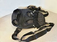 Front view of black breathable mesh pet backpack with visible openings for limbs and head, adjustable straps laid out, and zipper detail on top.