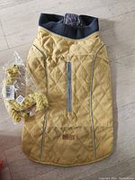 Yellow quilted dog coat laid flat showing reflective strip on back, ribbed collar, and chest Velcro. Yellow giraffe chew toy visible in packaging.