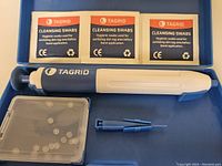 Open box showing TAGRID skin tag remover device, three cleansing swabs, a case with 40 rubber removal bands, and two blue applicator tools inside a blue plastic case.