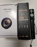 Side-by-side photo of MWC200 webcam box and Belike USB flashlight box and flashlight showing branding.
