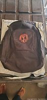 Full front view of black backpack with front pocket showing pink embroidered monogram letters.