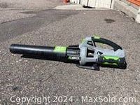 Side view of electric cordless handheld leaf blower sitting on asphalt surface with black blower tube and gray-green handle.