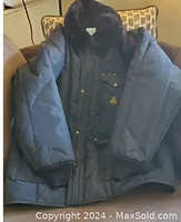 Front view of men's black Refrigiwear ski jacket with faux fur collar and snap-closure front pockets, displayed on a chair.