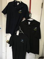 Two polo shirts (navy blue and black) and one t-shirt (black), all hanging on hangers against a white door with visible embroidered logos on left chest.