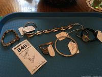 Watch with black leather strap and five bracelets arranged on a blue tray.