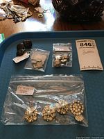 Various sets of buttons displayed on a blue tray, including a large plastic bag containing five gold-tone beaded floral cluster buttons, smaller bags with different styles of buttons, and two heart-shaped dark buttons with tags.