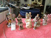 Overview of 11 Victorian-style ceramic figurines and 2 lamp bases on round table with pink tablecloth