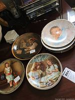 Photo of eight printed collector plates with various scenes including children, two girls having tea, a boy at a desk, and baby face design.
