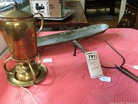 Photo of brass coffee warmer, copper drip pan, and pie lifter on pink fabric surface with lot tag.