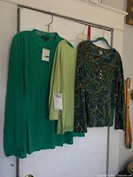 Set of three women's tops hanging on hangers on a wall. Left is a bright green embellished top with new tag (Greater Good). Middle is lime green Coldwater Creek top with scalloped sleeve edges. Right is darker paisley patterned embellished Chicos top.