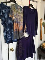 Four ladies tops hanging on clothes hangers against a white door: blue wash frilly short sleeve top, glittery patterned short sleeve top, plain purple long sleeve top, and purple 3/4 sleeve top with gold accent pattern.
